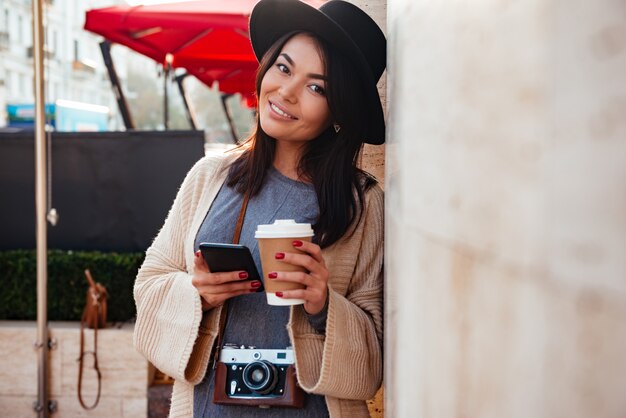 Vrolijke Aziatische vrouw in stijlvolle smartphone van de slijtageholding en meeneemkoffie, die camera bekijken, terwijl status op de stadsstraat