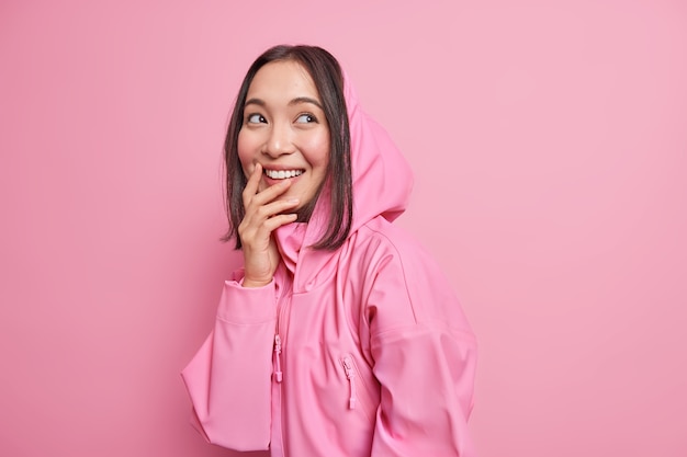 Vrolijke Aziatische vrouwelijke tiener staart dagdromen opzij en herinnert zich een mooi moment glimlacht breed gekleed in hoodie drukt positieve openhartige emoties uit geïsoleerd over roze muur. Jeugd levensstijl