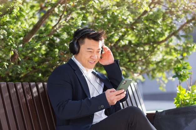 Vrolijke aziatische zakenman zittend op een bankje in een stadspark in het centrum en luisteren naar muziek op koptelefoon met telefoon in zijn handen. man in een pak geniet van het buitenleven. ontspan en rust. levensstijl