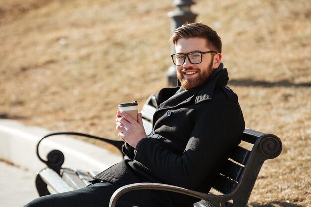 Gratis foto vrolijke bebaarde jonge man zittend op een bankje en koffie drinken