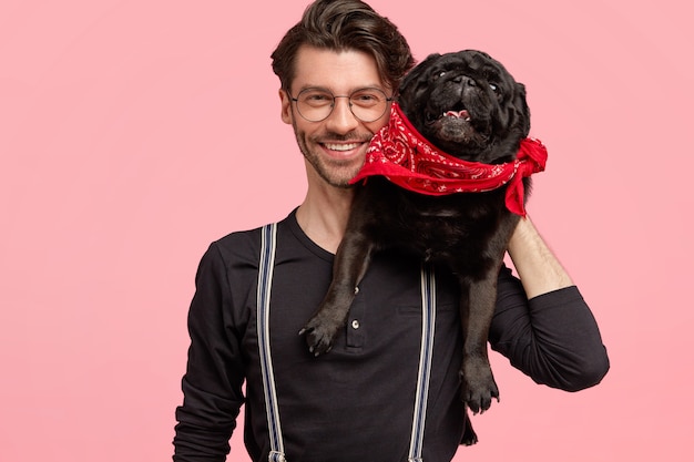 Gratis foto vrolijke bebaarde man heeft brede glimlach, blij om te poseren met zijn rashond, houdt van huisdieren, gekleed in modieus zwart shirt en bretels, geïsoleerd over roze muur. gelukkig man met dier