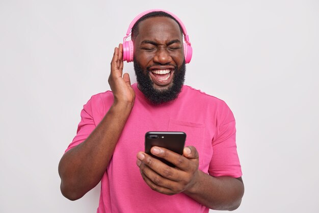 Vrolijke bebaarde man met geniet van audiotrack in koptelefoon maakt gebruik van moderne technologieën luistert favoriete nummer besteedt vrije tijd aan hobby draagt casual t-shirt geïsoleerd over grijze muur