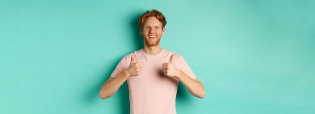 Gratis foto vrolijke bebaarde man met rood haar die duim omhoog laat zien en iets goedkeurt dat promo standi prijst