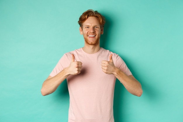 Vrolijke bebaarde man met rood haar die duimen omhoog laat zien, iets leuk vindt en goedkeurt, promo prees, staande tegen een turkooizen achtergrond