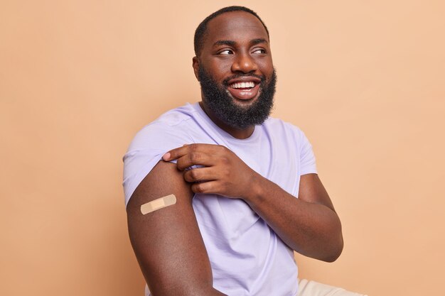 Vrolijke bebaarde man toont schouder met plakband na vaccinatie ontvangen coronavaccin beige muur
