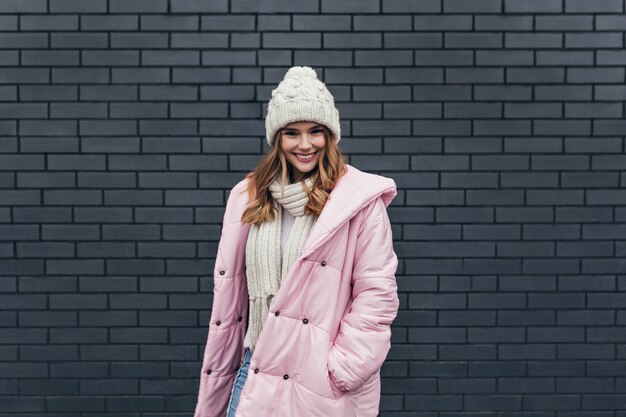 Vrolijke blanke dame draagt roze jas plezier in koude dag. Buiten foto van vrolijk meisje permanent in de buurt van dichtgemetselde muur en glimlachen.