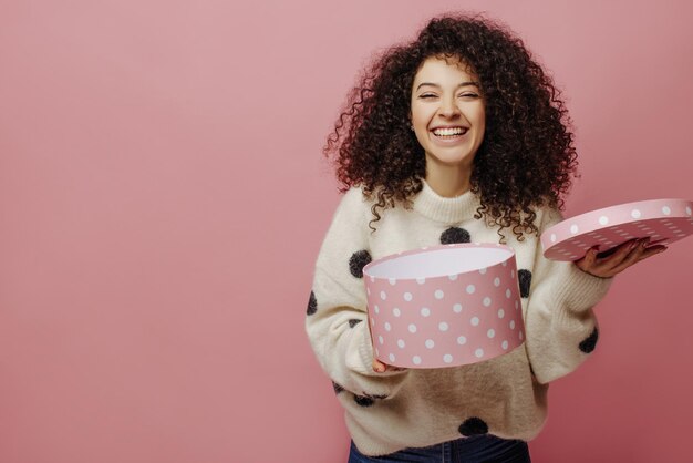 Vrolijke blanke jonge dame verheugt zich over de ontvangen geschenkdoos in handen op roze achtergrond Meisje met zwart krullend haar draagt witte trui Concept van vieringsgeluk en emoties