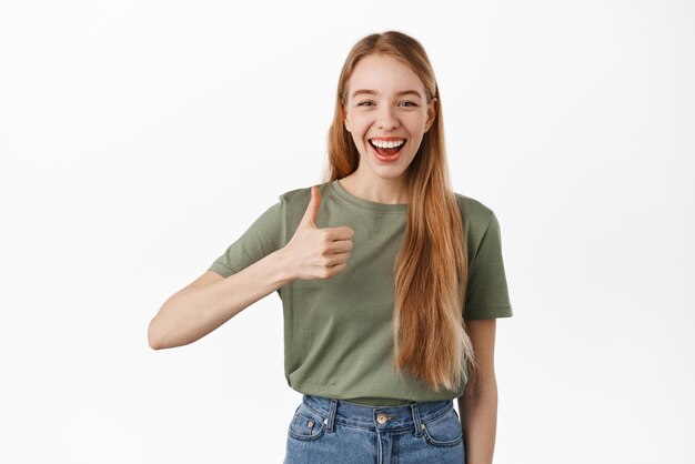 Vrolijke blanke vrouw toont duimen en zegt ja glimlachend tevreden beveel bedrijf of merk aan, keur goed en houd van iets goeds lof keuze staande op een witte achtergrond