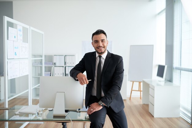 Vrolijke blanke zakenman zittend op een bureau in het kantoor, leunend op het scherm en glimlachen