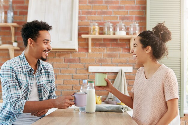 Vrolijke blije vrouwen en mannen in vrijetijdskleding drinken melk in de keuken, lachen luid terwijl ze elkaar grappige verhalen vertellen.