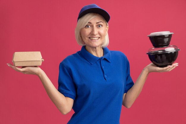 Vrolijke blonde bezorger van middelbare leeftijd in blauw uniform en pet met papieren voedselpakket en voedselcontainers