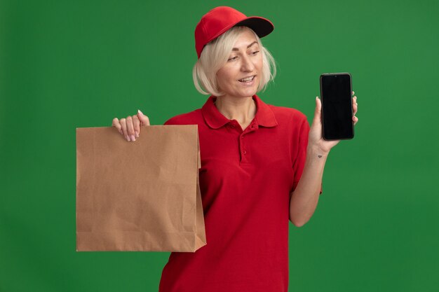 Vrolijke blonde bezorger van middelbare leeftijd in rood uniform en pet met papieren pakket en mobiele telefoon die naar de telefoon kijkt