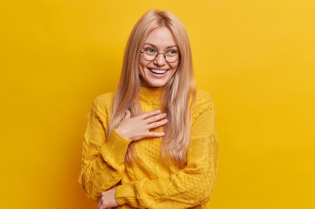 Vrolijke blonde Europese vrouw lacht vrolijk kijkt zorgeloos giechelt positief hand op de borst houdt
