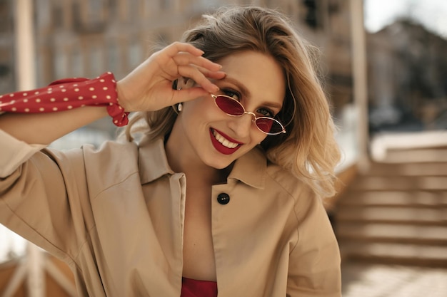 Vrolijke blonde vrouw in een goed humeur zet een heldere zonnebril op Gelukkige dame met rode lippen gekleed in beige trenchcoat glimlacht naar buiten