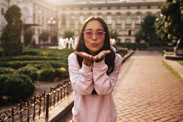 Vrolijke brunette dame in kleurrijke zonnebril en roze hoodie poseert in een goed humeur buiten en blaast kus