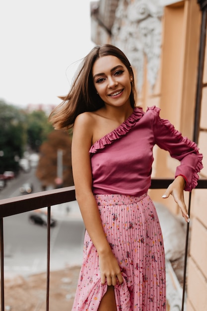 Vrolijke brunette met kort haar mooie glimlach. Gelukkig vrouwelijk model in roze blouse beweegt op balkon