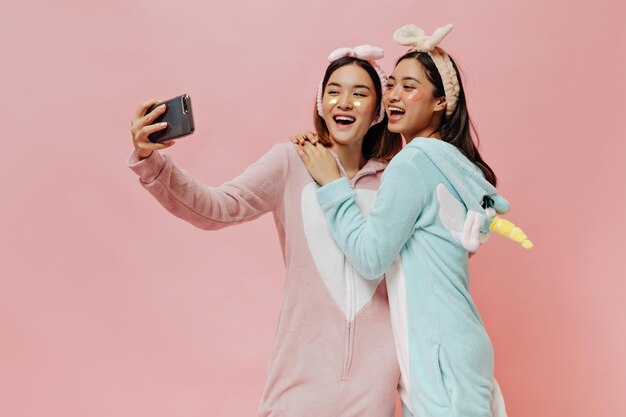 Vrolijke brunette vrouwen in kigurumi's en hoofdbanden glimlachen oprecht en nemen selfie op roze geïsoleerde achtergrond