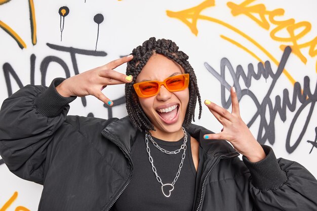 Vrolijke coole etnische vrouw met dreadlocks maakt yo gebaar heeft plezier gekleed in zwarte jas en stijlvolle oranje zonnebril glimlacht breed poseert tegen graffiti muur