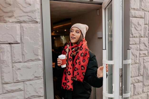 Vrolijke dame in beige pet en rode sjaal komt uit cafetaria met koffie in zwarte jas Outdoor portret van blij jonge vrouw rusten in restaurant op straat
