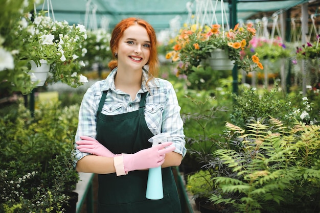 Vrolijke dame in schort en roze handschoenen die met een spuitfles in de hand staan en vrolijk in de camera kijken terwijl ze in de kas werkt