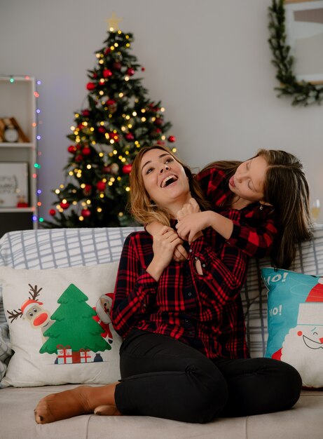 Vrolijke dochter stak tong en knuffels moeder zittend op de Bank van achteren genieten van kersttijd thuis