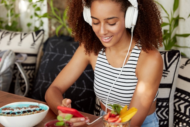 Vrolijke donkere vrouw controleert e-mailbox online op slimme telefoon, verbonden met draadloos internet bij coffeeshop, luistert coole populaire muziek in koptelefoon, downloadt audioboek, eet dessert