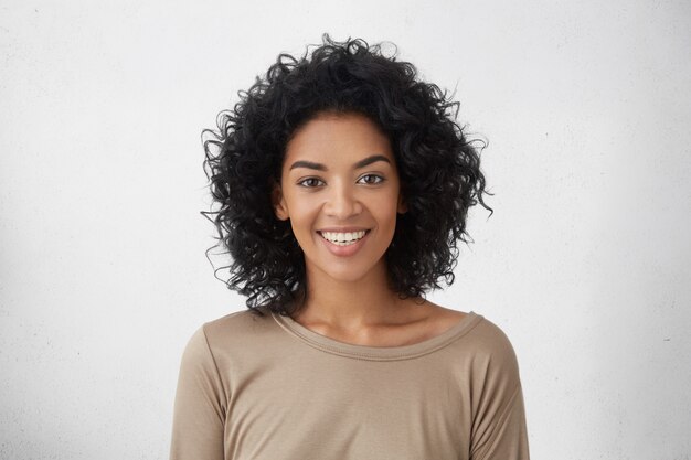 Vrolijke donkere vrouw die breed glimlacht, zich verheugt over haar overwinning in concurrentie tussen jonge schrijvers, staande geïsoleerd tegen de grijze muur. Mensen, succes, jeugd en geluk concept