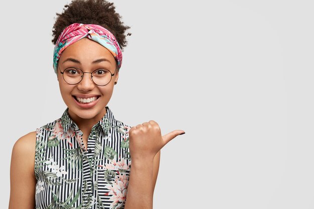 Vrolijke donkere vrouw heeft een vriendelijke glimlach, gekleed in blouse met bloemenprint, wijzend op copyspace