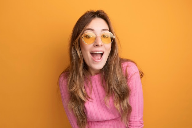 Vrolijke en gelukkige jonge mooie vrouw in roze top met een bril die naar een camera kijkt die lacht over een oranje achtergrond