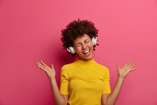 Vrolijke, geamuseerde, donkere, gekrulde jonge vrouw geniet van geweldige koptelefoonkwaliteit, prachtig geluid, handpalmen omhoog, luistert naar muziek, voelt zich tevreden met een goed lied, gekleed in gele kleren, heeft plezier