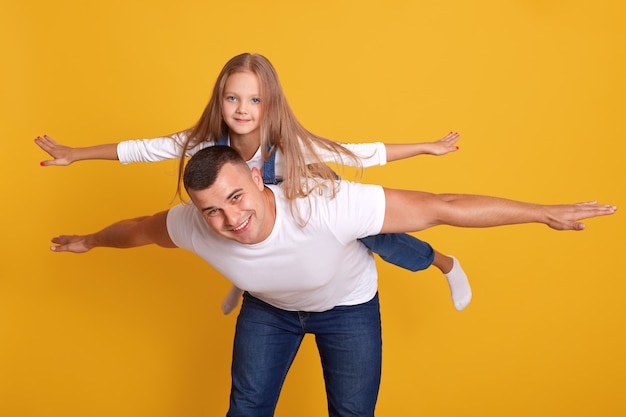 Vrolijke gelukkig man verbeeldt zich met zijn mooie dochter om te vliegen als een vliegtuig, poseren geïsoleerd op geel. schot, gelukkige momenten met de beste vader, saamhorigheid, familieconcept.