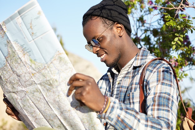 Vrolijke gelukkige jonge afro-amerikaanse reiziger met een trendy look die op zoek is naar een richting op de locatiekaart, op zoek naar het hotel terwijl hij naar het buitenland reist in een buitenlandse stad tijdens de zomervakantie