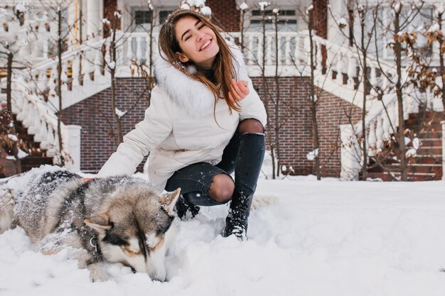 Vrolijke gelukkige jonge vrouw met plezier met schattige husky hond in de sneeuw op straat. Vrolijke stemming, wintersneeuwtijd, lieve huisdieren, echte vriendschap.