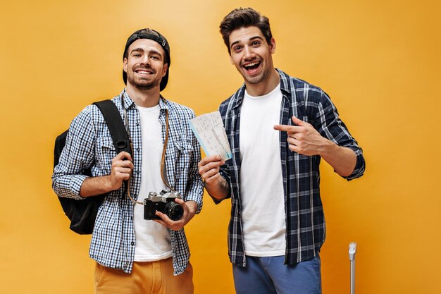 Vrolijke gelukkige reizigers poseren en verheugen zich op een oranje achtergrond Jonge mannen in geruite overhemden houden vliegtickets en retro camera vast