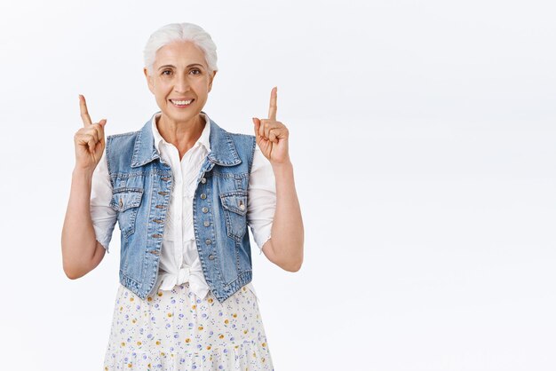 Vrolijke glimlachende oude kaukasische vrouw in denim vest over jurk grijnzend gelukkig wijzende vingers omhoog aanbevelen product suggereren nuttige link trots tonen promo staande witte achtergrond