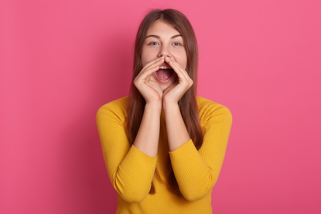 Vrolijke grappige jonge vrouw, gekleed in gele trui schreeuwen met handgebaar in de buurt van mond geïsoleerd over roze muur, dame met lang haar