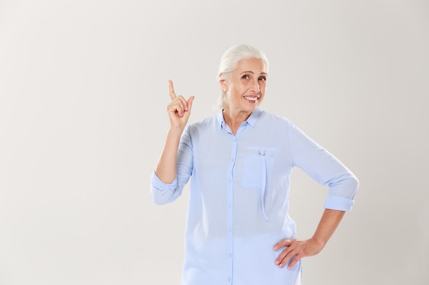Vrolijke grijze oude dame in blauw shirt, wijzend met de vinger naar boven