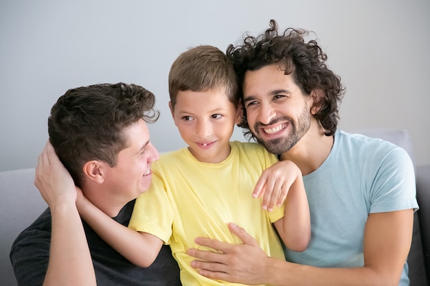 Vrolijke homo vaders en zoon samen op de bank zitten en elkaar knuffelen. Vooraanzicht. Gelukkig gezin en ouderschap concept