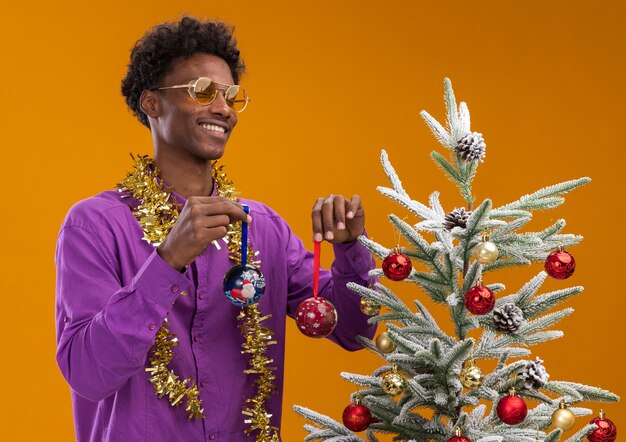 Vrolijke jonge Afro-Amerikaanse man met bril met klatergoud slinger rond nek staande in de buurt van versierde kerstboom op oranje achtergrond