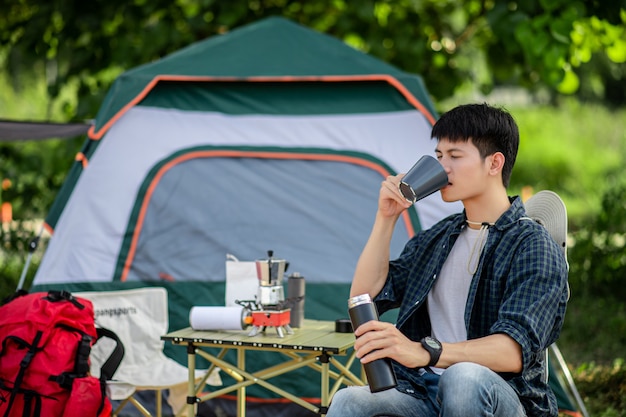 Vrolijke jonge backpacker man zit aan de voorkant van de tent in het bos met koffieset en het maken van verse koffiemolen tijdens het kamperen op zomervakantie