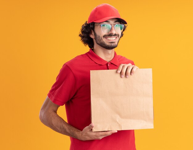 Vrolijke jonge bezorger in rood uniform en pet met een bril die een papieren pakket vasthoudt en kijkt naar de voorkant geïsoleerd op een oranje muur
