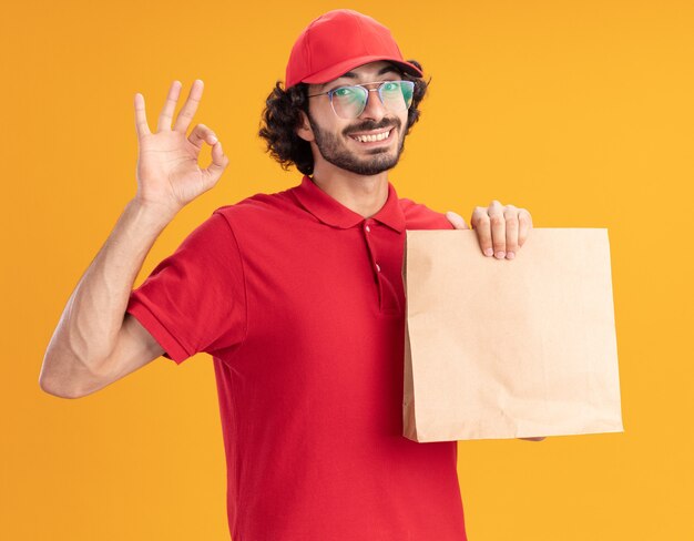 Vrolijke jonge bezorger in rood uniform en pet met een bril die een papieren pakket vasthoudt en naar de voorkant kijkt met een ok teken geïsoleerd op een oranje muur orange