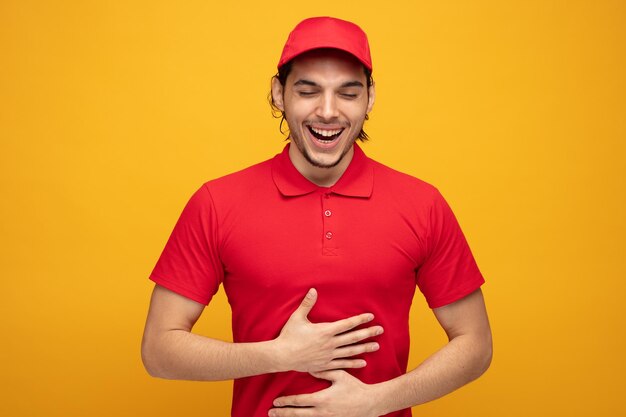 Vrolijke jonge bezorger met uniform en pet lachend met gesloten ogen terwijl hij zijn handen op zijn buik houdt geïsoleerd op gele achtergrond