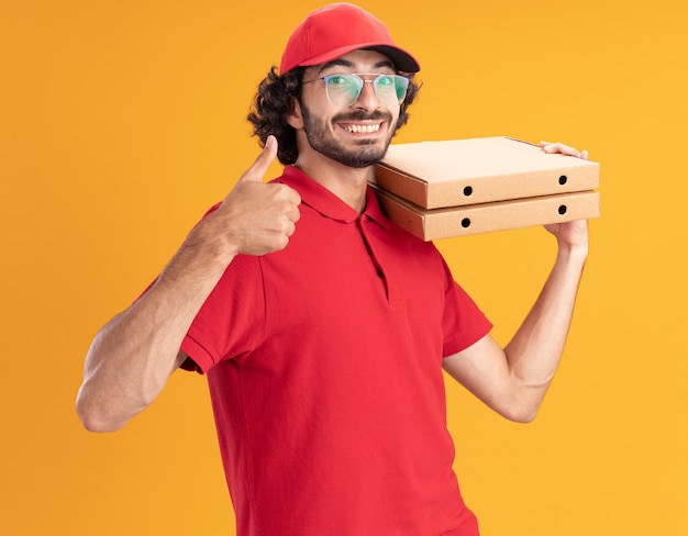 Vrolijke jonge blanke bezorger in rood uniform en pet met een bril die in profielweergave staat en pizzapakketten op de schouder houdt met duim omhoog geïsoleerd op oranje muur