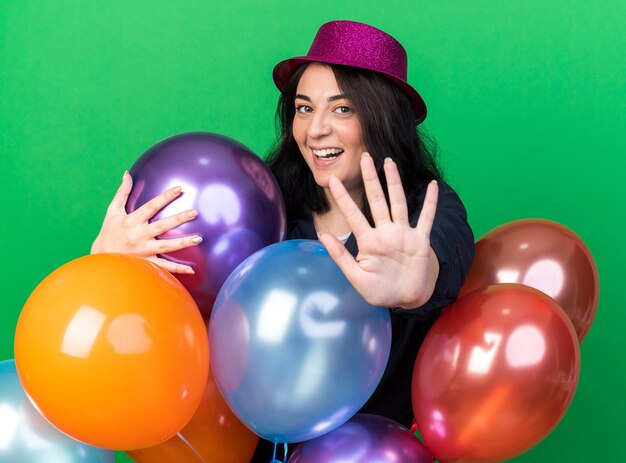 Vrolijke jonge blanke feestvrouw met een feesthoed die achter ballonnen staat en ze knuffelt terwijl ze naar de voorkant kijkt en een stopgebaar doet geïsoleerd op een groene muur