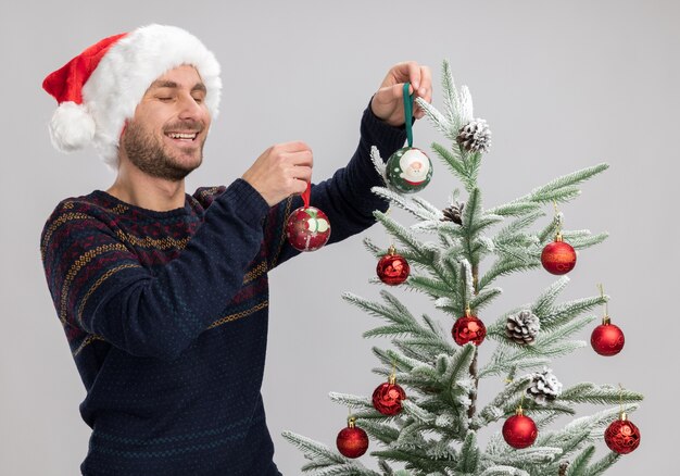 Vrolijke jonge blanke man met kerstmuts staande in de buurt van de kerstboom en versiert het met kerst ornament ballen lachen met gesloten ogen geïsoleerd op een witte achtergrond