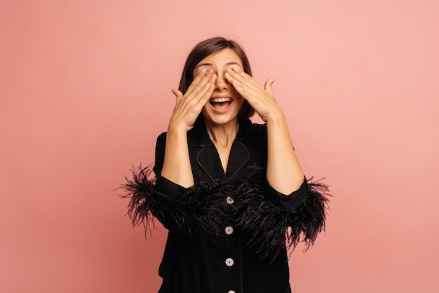 Vrolijke jonge blanke meisje in zwart shirt met open mond sluit ogen met handen terwijl staande op roze achtergrond Emotie concept