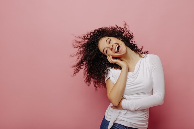 Vrolijke jonge blanke meisje lachen op de top van haar stem zwaaien krullend zwart haar op roze achtergrond Vrouw draagt witte blouse plezier vrije tijd emoties en lifestyle concept