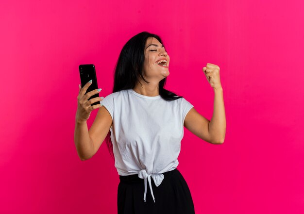 Vrolijke jonge blanke vrouw houdt telefoon vast en houdt haar vuist omhoog kijkend naar de zijkant