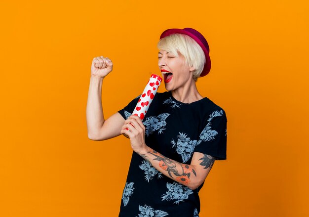 Vrolijke jonge blonde partij meisje met feestmuts zingen met behulp van confetti kanon als microfoon met gesloten ogen geïsoleerd op een oranje achtergrond met kopie ruimte